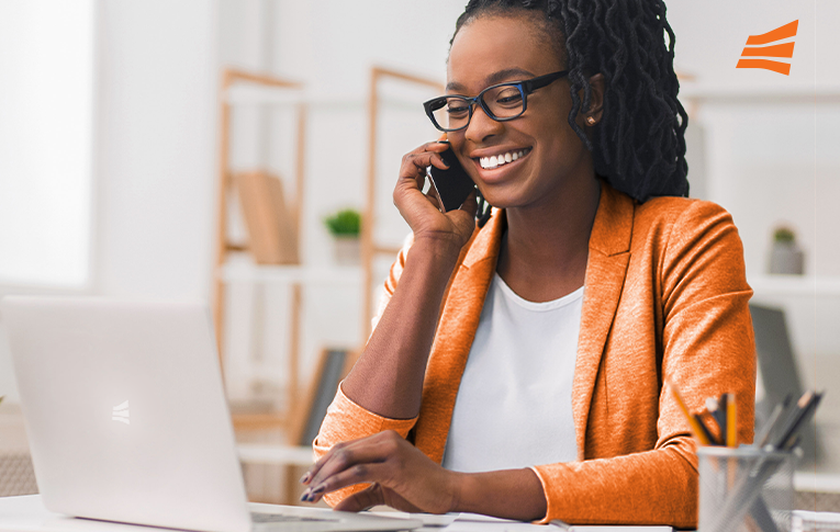 Na imagem: mulher negra, vestindo um blazer laranja, sorrindo enquanto usa um notebook e conversa com o cliente pelo celular, na estratégia de custommer success.