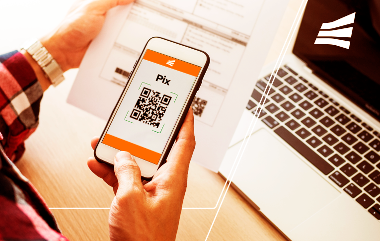 Na imagem: uma mão segurando o smartphone enquanto lê o QR Code de um papel impresso, para pagar com Pix.