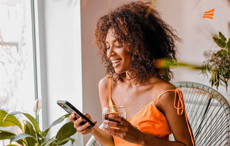 Mulher sorridente com uma xícara de café na mão e o smartphone na outra, usando sua Conta Digital da Gerencianet, na varanda de casa.