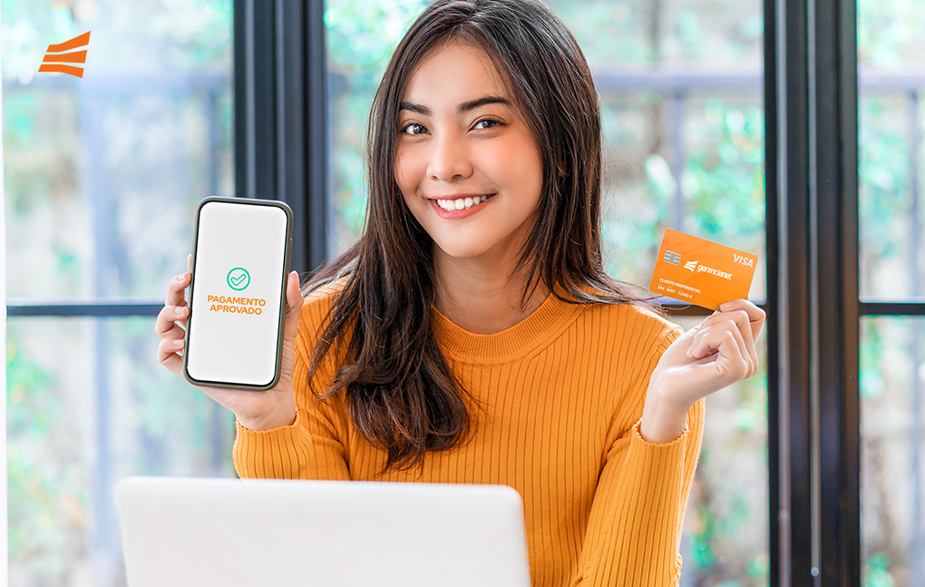 Mulher sorrindo em frente ao notebook, com o celular em uma mão e o Cartão de Crédito Gerencianet na outra, ilustrando que existem as duas maneiras de como pagar a fatura do cartão de crédito: web ou app.
